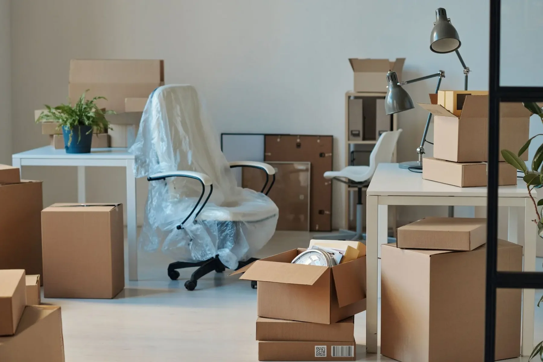 Office space filled with cardboard moving boxes, a desk lamp, and a chair covered in plastic wrap.