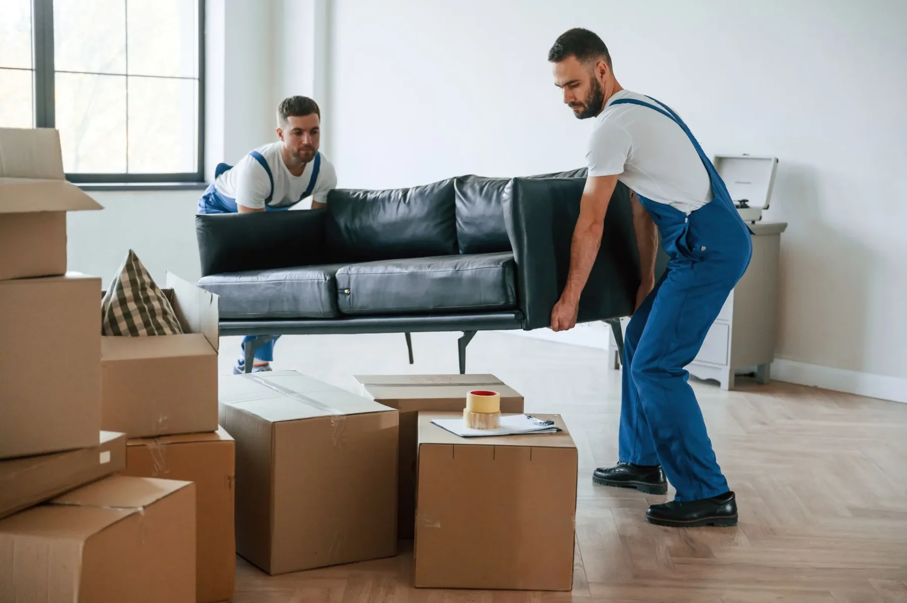 Two movers in blue overalls carrying a black sofa across a room filled with cardboard moving boxes.